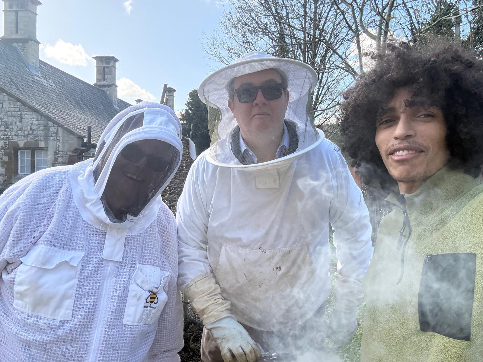 Two beekeepers in white suits and veils lifting a frame from an open hive, smoke drifting across the grass